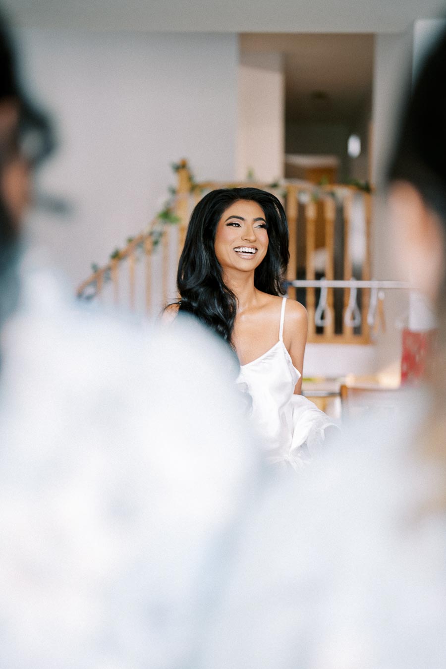 Young woman in a white dress smiling brightly indoors, with a floral-decorated staircase in the background.