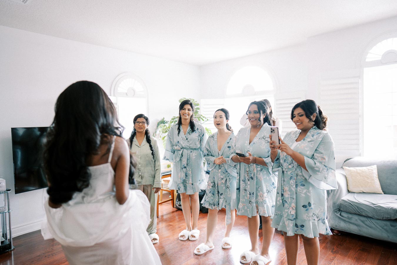 A bride wearing a white dress is seen with her back to the camera, facing five bridesmaids in matching light blue floral robes and slippers. The bridesmaids are smiling and holding phones, capturing a joyful pre-wedding moment in a sunlit living room with wooden floors and a couch.