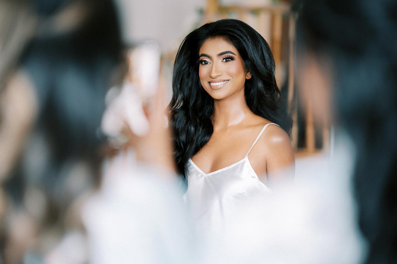 Portrait of a smiling woman with long black hair wearing a white silk tank top, captured in a soft-focus setting.