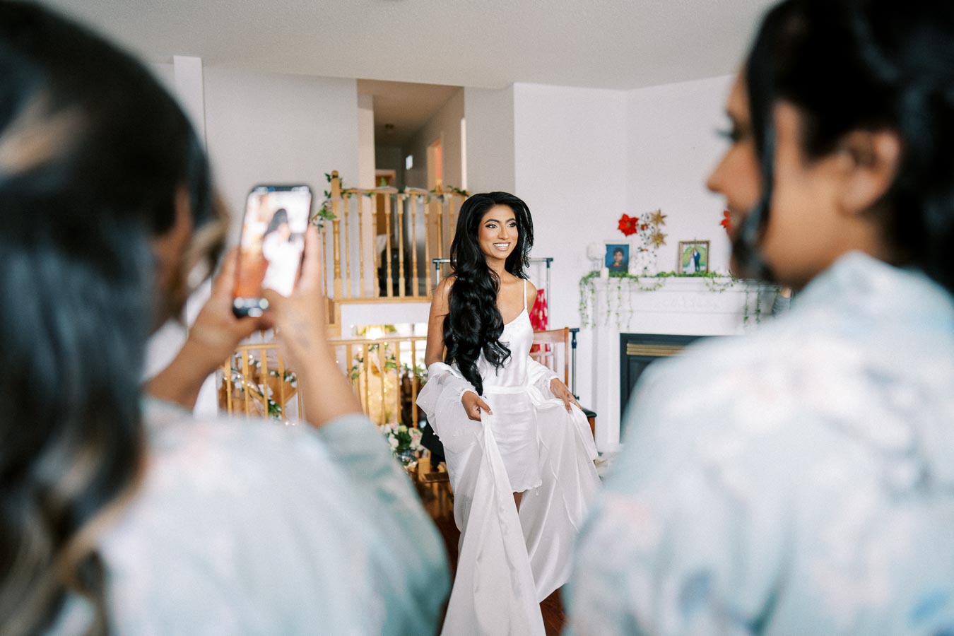 A bride in a white robe smiles while getting ready for her wedding, surrounded by bridesmaids capturing the moment on their phones in a beautifully decorated living room.