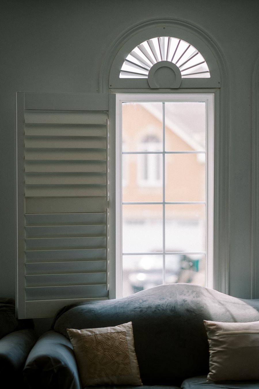 Elegant living room with a classic arched window featuring white shutters, a gray sofa, and decorative pillows, creating a cozy interior ambiance.