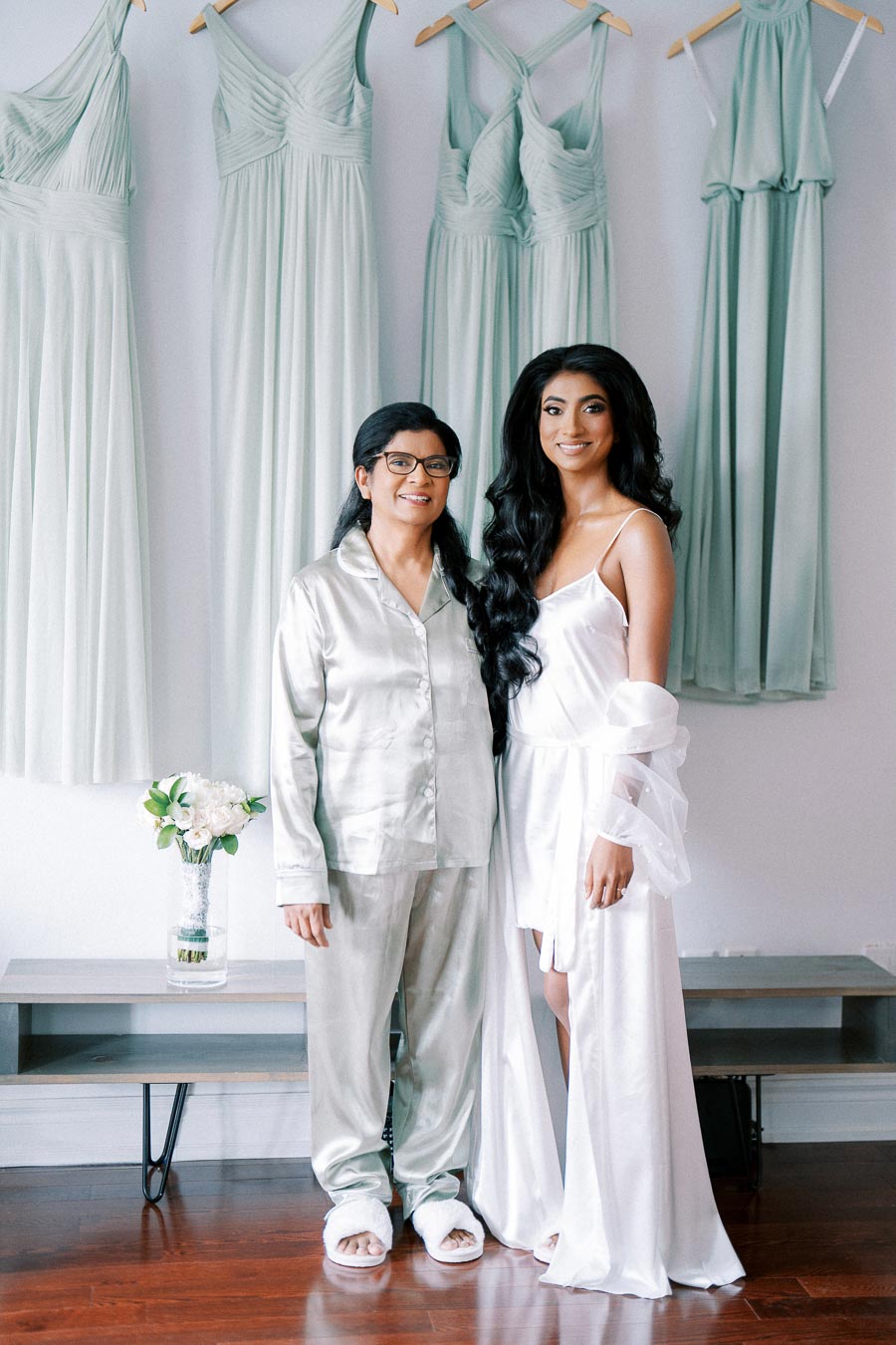 Two women smiling in a bridal preparation setting, wearing elegant satin robes, with pastel bridesmaid dresses hanging in the background.
