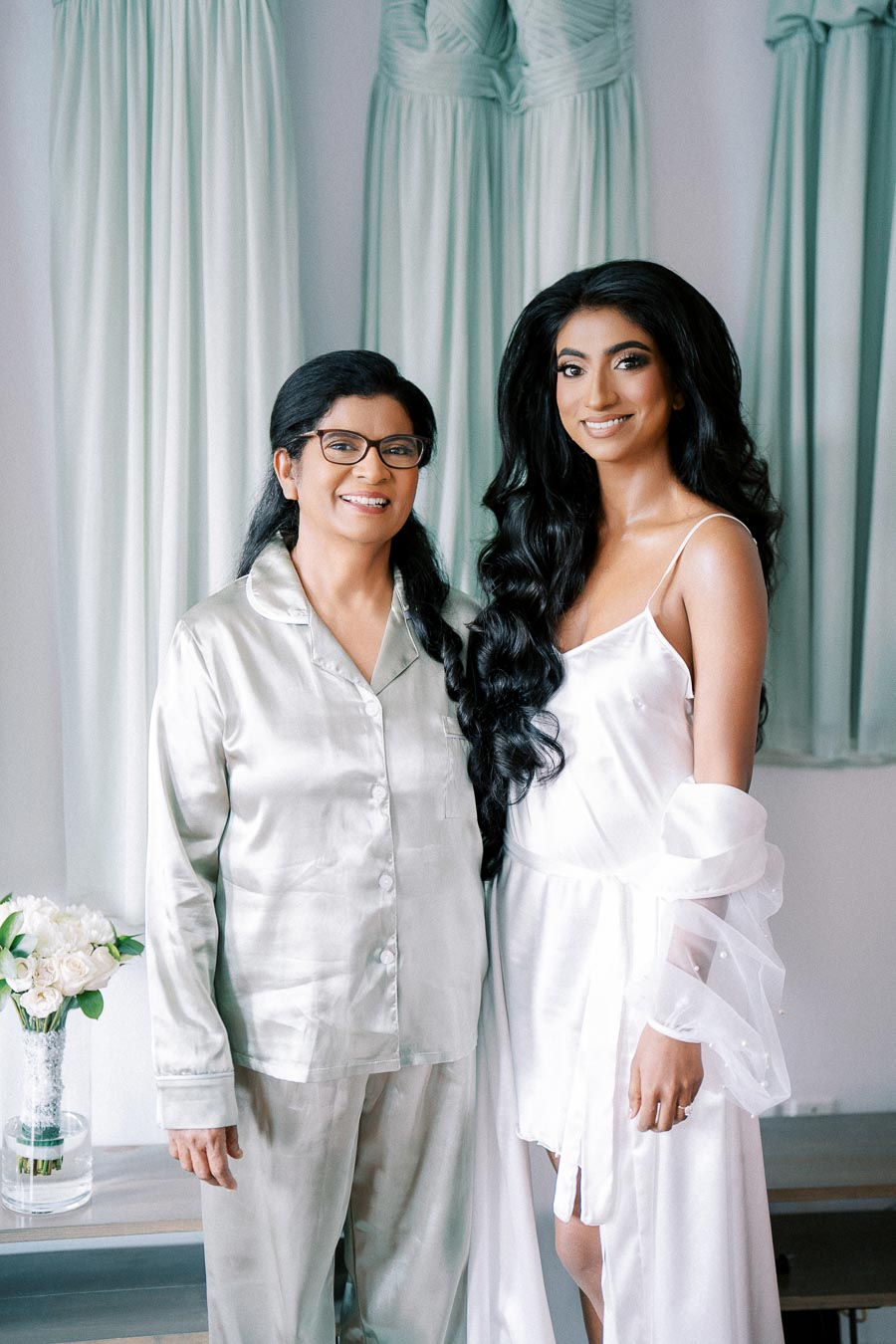 A bride and her mother smiling, standing together in front of hanging pastel turquoise dresses, with the bride in a white silk robe and her mother in silver satin attire, holding a bouquet of white flowers in the background.