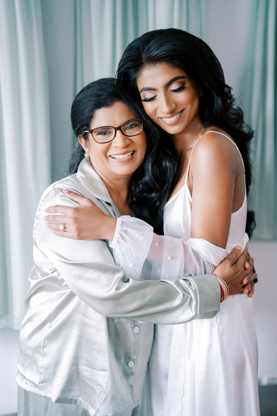 A bride and her mother share an affectionate moment, embracing lovingly in preparation for the wedding. The bride's hair is elegantly styled, and she wears a white satin dress, while her mother wears glasses and a silk outfit. Light curtains serve as a soft backdrop, enhancing the intimate and joyful scene.