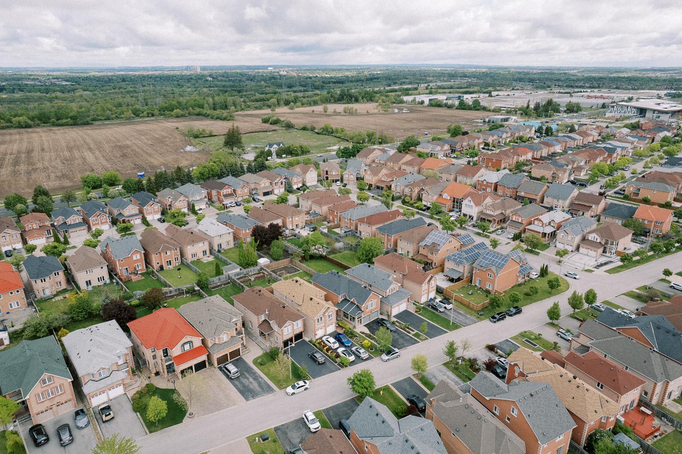 Aerial view of a suburban neighborhood with rows of houses featuring different colored roofs, surrounded by green lawns and trees, under a cloudy sky. A large field and additional greenery are visible in the background.
