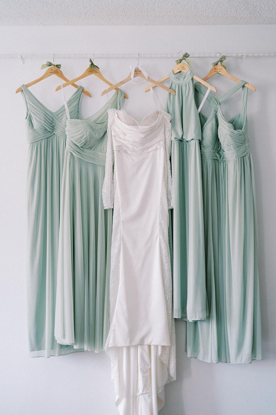Elegant bridal gown and bridesmaid dresses on display, featuring a white wedding dress and five soft green gowns hanging on wooden hangers, ideal for a stylish wedding ensemble inspiration.