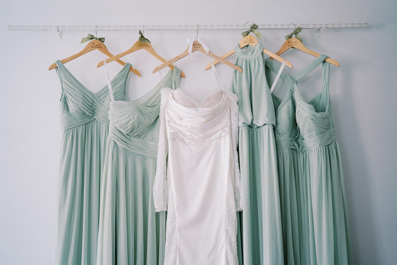 Five elegant dresses hanging on a rack, featuring a central white bridal gown surrounded by four sage green bridesmaid dresses, perfect for a stylish wedding ensemble.