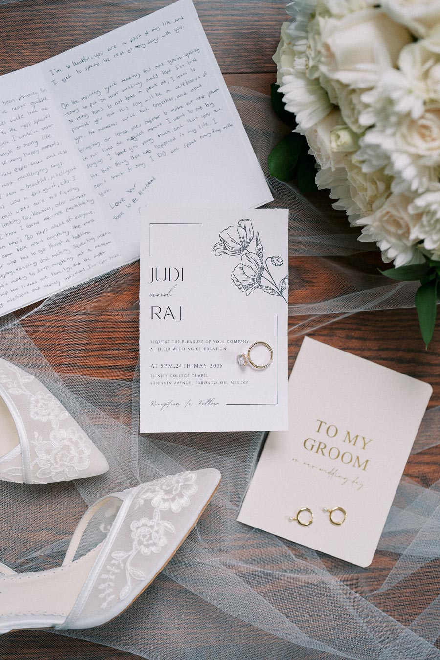 Wedding invitation flat lay with Judi and Raj's invitation card featuring floral design, elegant handwritten letter, white bridal shoes with lace embroidery, gold ring, and white bouquet on wooden background.