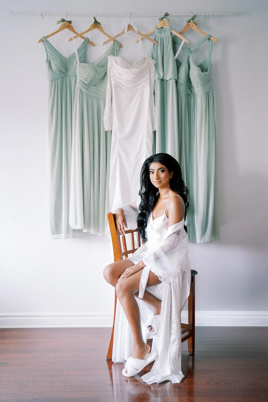A bride in a white satin robe sits on a wooden chair, looking away from a rack of hanging mint green bridesmaid dresses and a white wedding gown against a minimalistic background.