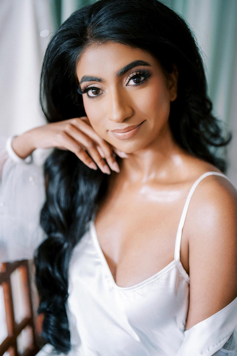 Elegant woman with long dark hair and natural makeup wearing a white satin dress, posing with hand on chin against a soft, blurred background