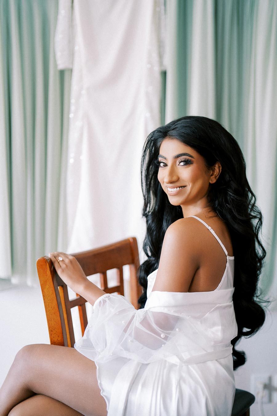 Bridal preparation with a smiling woman in a white robe, seated in front of a hanging wedding dress, elegant and joyful.