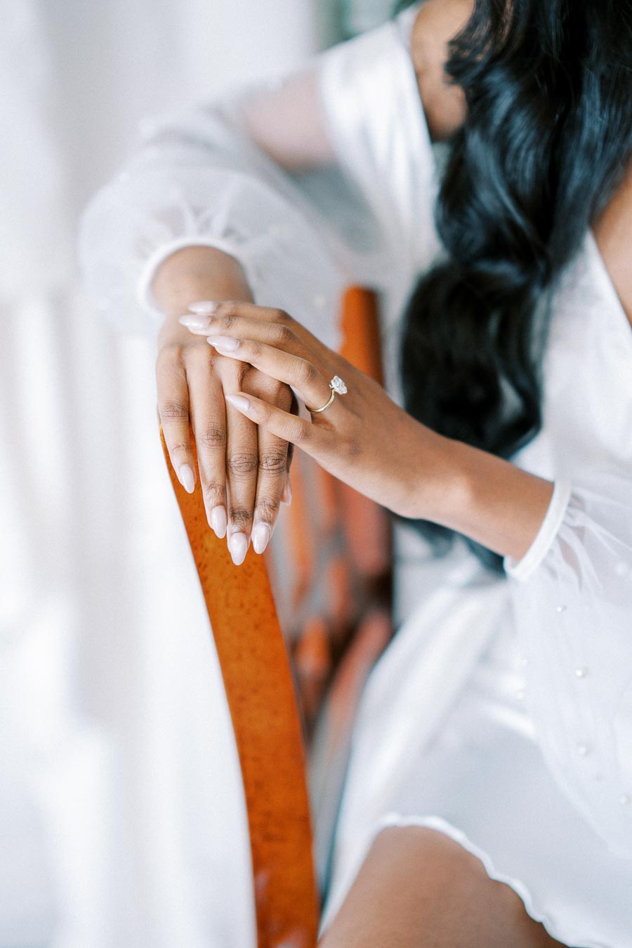 Close-up of a woman wearing an elegant engagement ring, showcasing manicured hands resting on a chair, with a soft-focus background of a white robe and long dark hair.