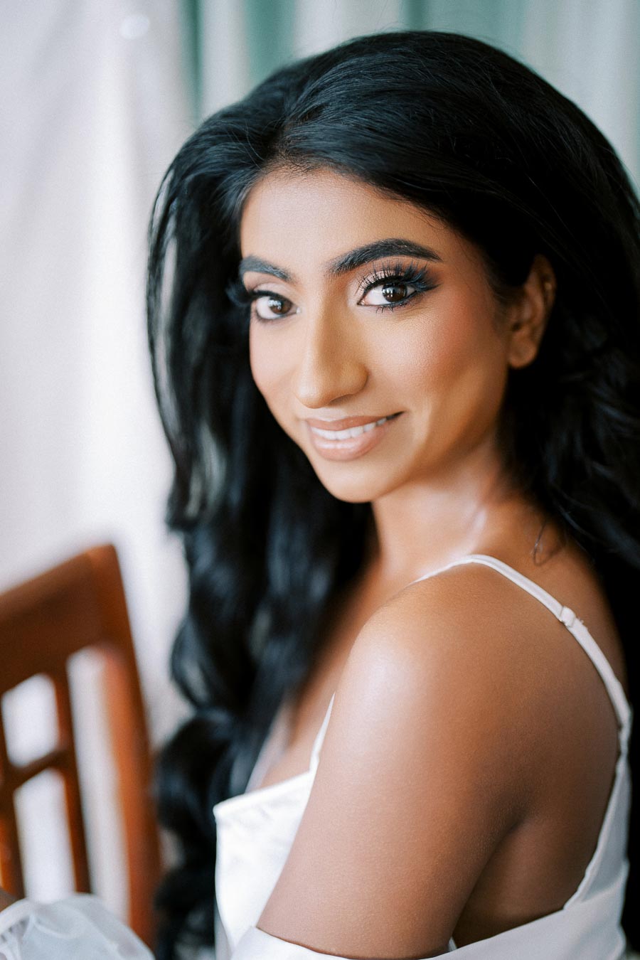 Smiling woman with long black hair and natural makeup wearing a white sleeveless top, sitting on a wooden chair with a soft focus background.