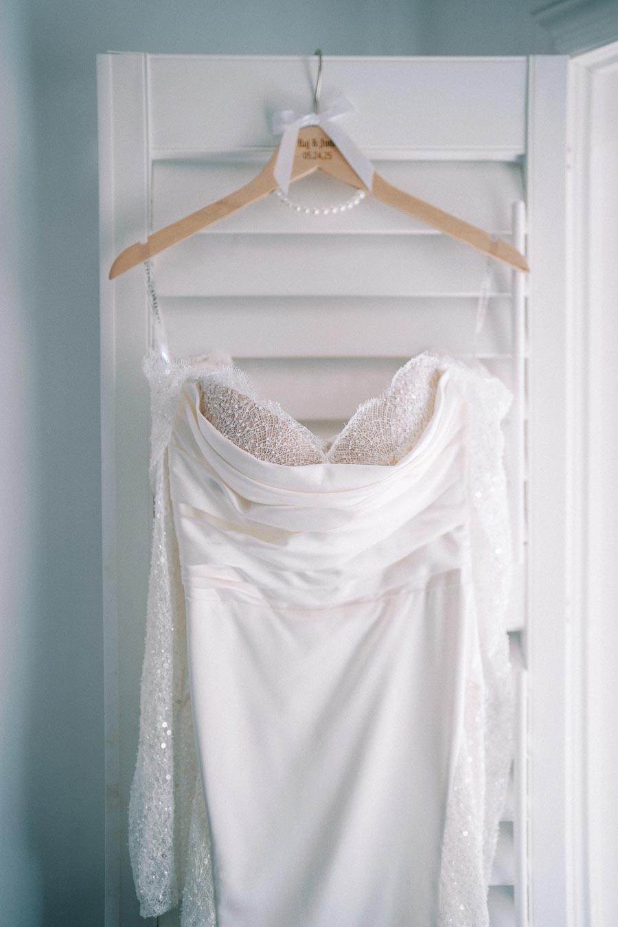 Elegant white wedding dress with lace detailing, hanging on a wooden hanger in front of white shutters, perfect for a bridal fashion look.