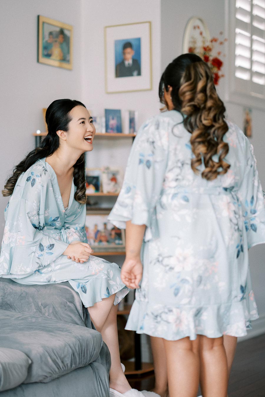Two women in floral robes sharing a joyful moment in a cozy room with framed photos and soft lighting.