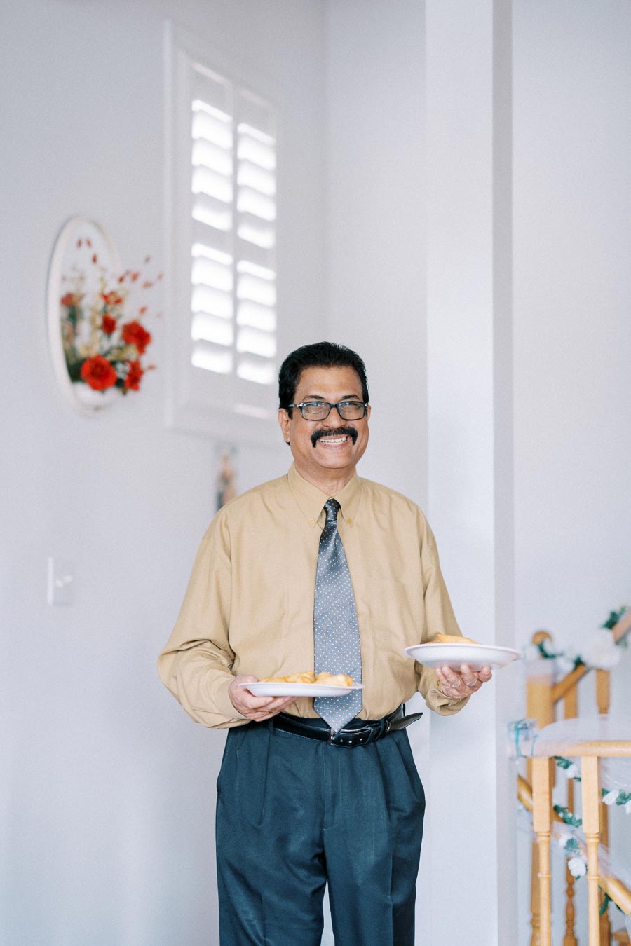 Man in a beige shirt smiling and holding plates of pastries, standing in a well-lit room with floral wall decor.