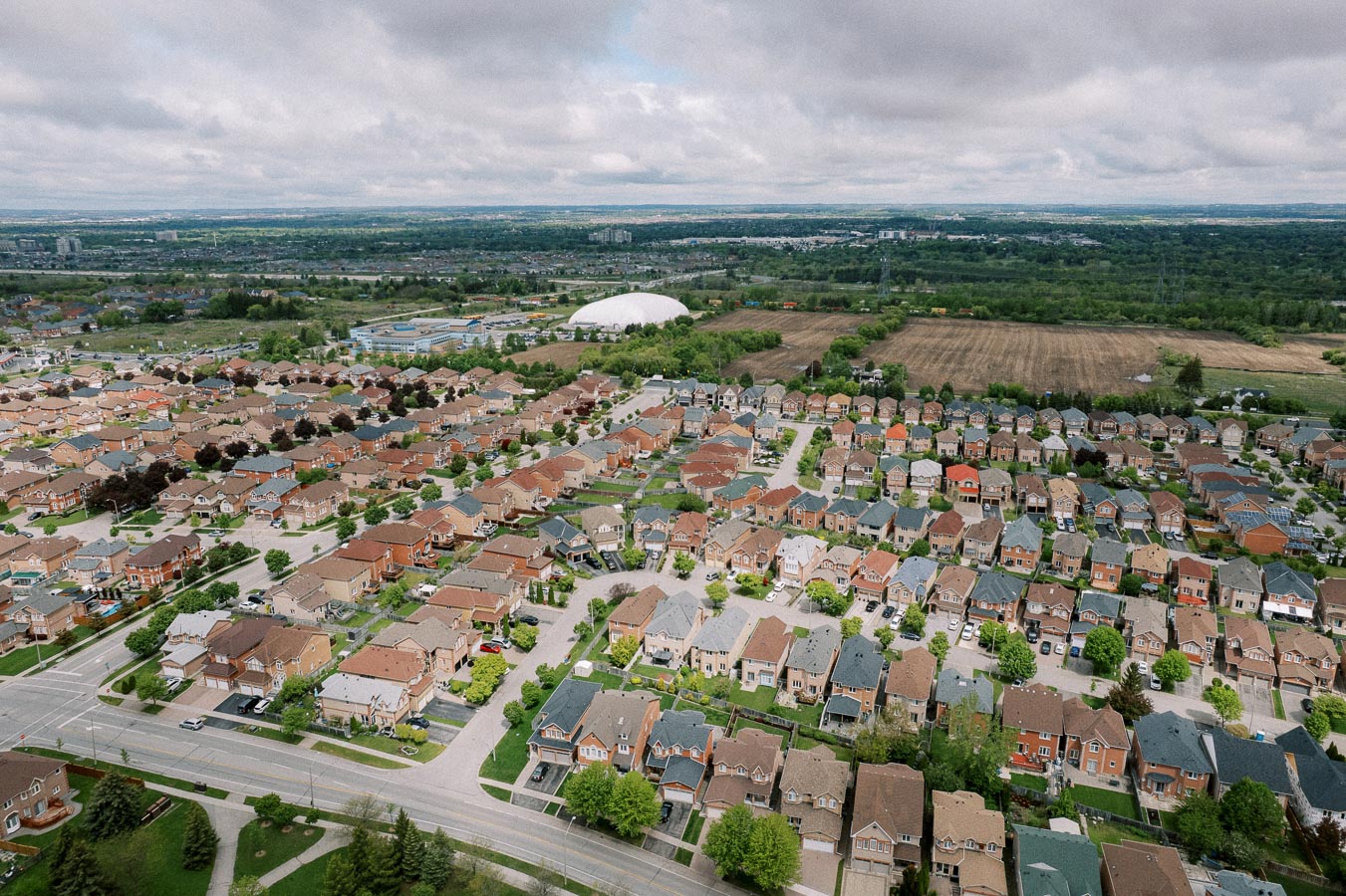 Aerial view of a suburban neighborhood with rows of houses featuring varied roof colors, surrounded by lush greenery and open fields under a cloudy sky.