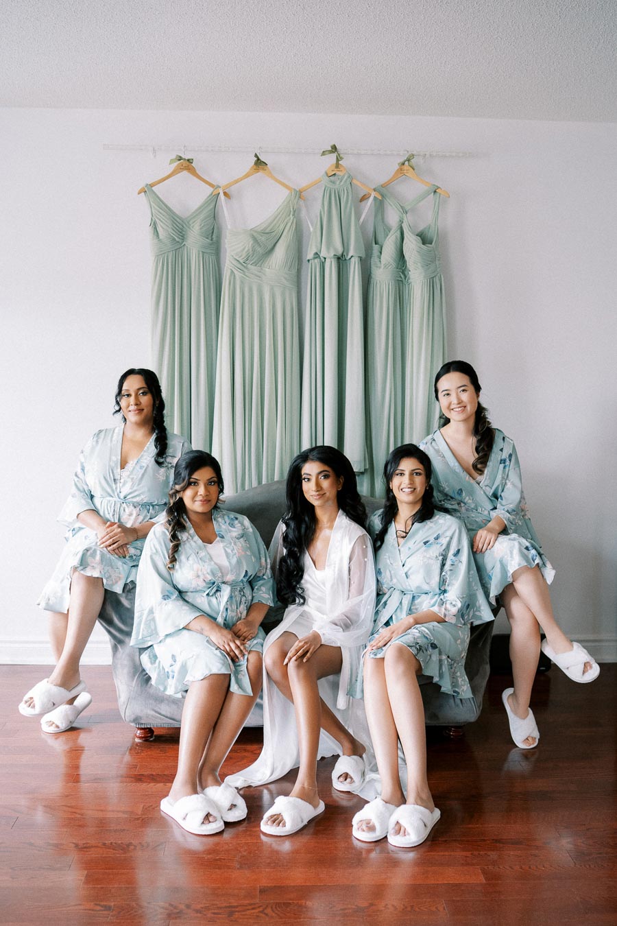 Bridesmaids sit together in matching floral robes and slippers, posing for a pre-wedding photo with bridesmaid dresses hanging in the background.