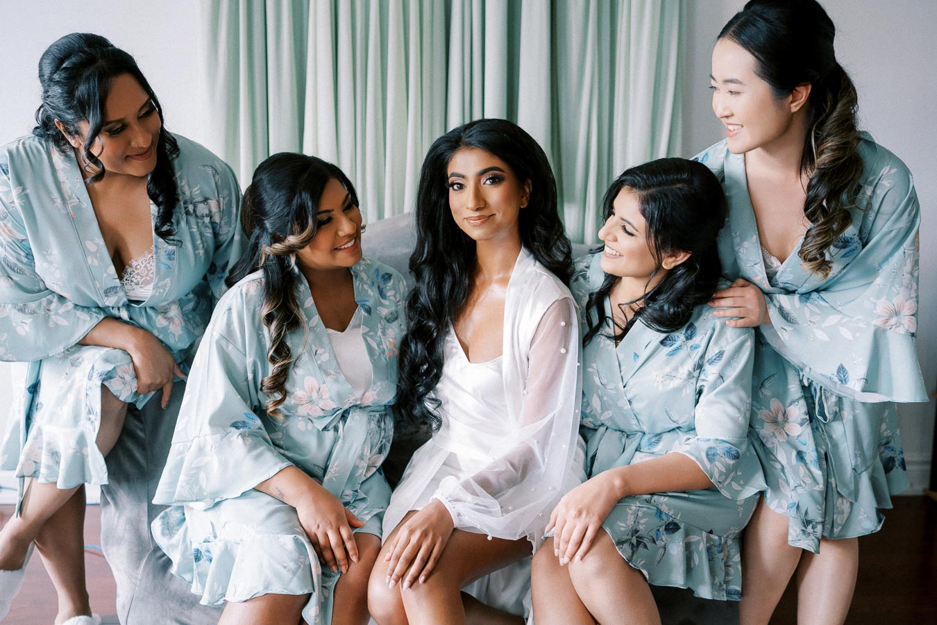 A bride surrounded by her bridesmaids wearing matching floral robes, preparing for a wedding day celebration.