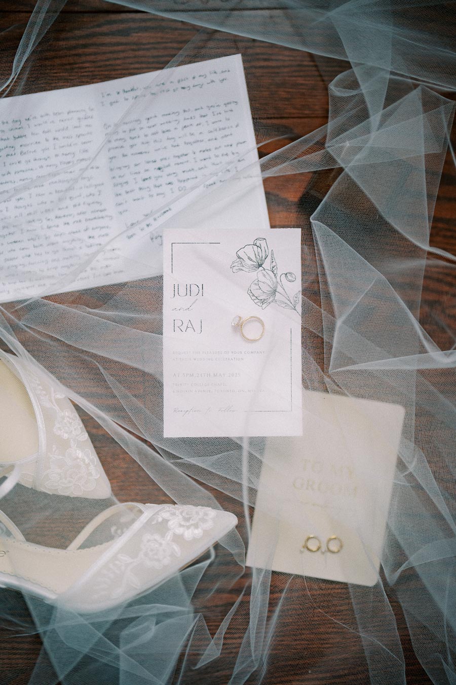 Elegant flat lay of wedding details on a wooden surface, featuring a bridal shoe with lace detailing, a handwritten letter, an invitation card with floral illustration, and wedding rings, all surrounded by delicate tulle fabric.