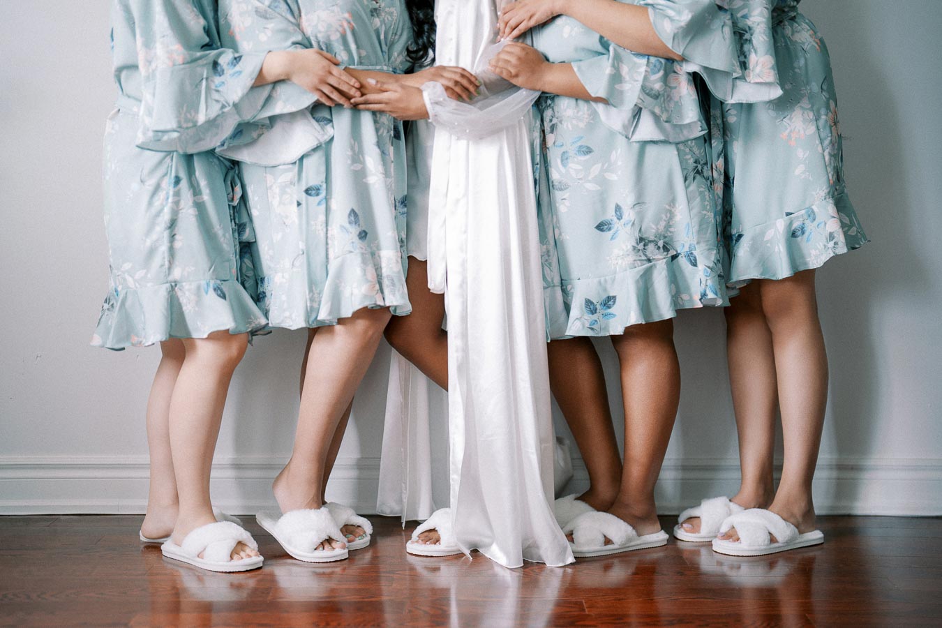 Bridal party in light blue floral robes and white slippers, standing together on a hardwood floor, sharing a special wedding day moment.