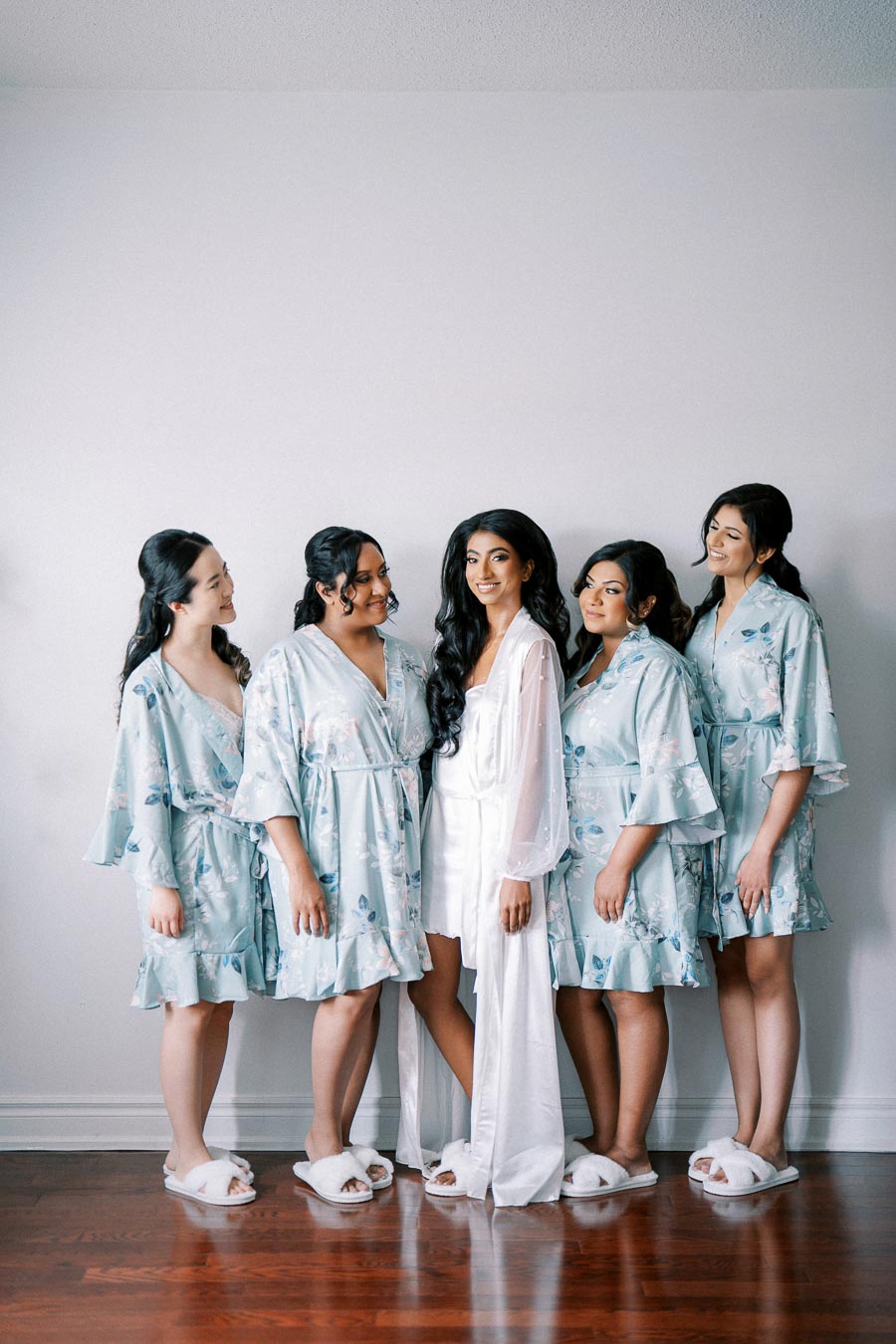 Bridal party posing in matching floral robes before the wedding, with the bride in a white robe and slippers, smiling with bridesmaids against a minimalist background.