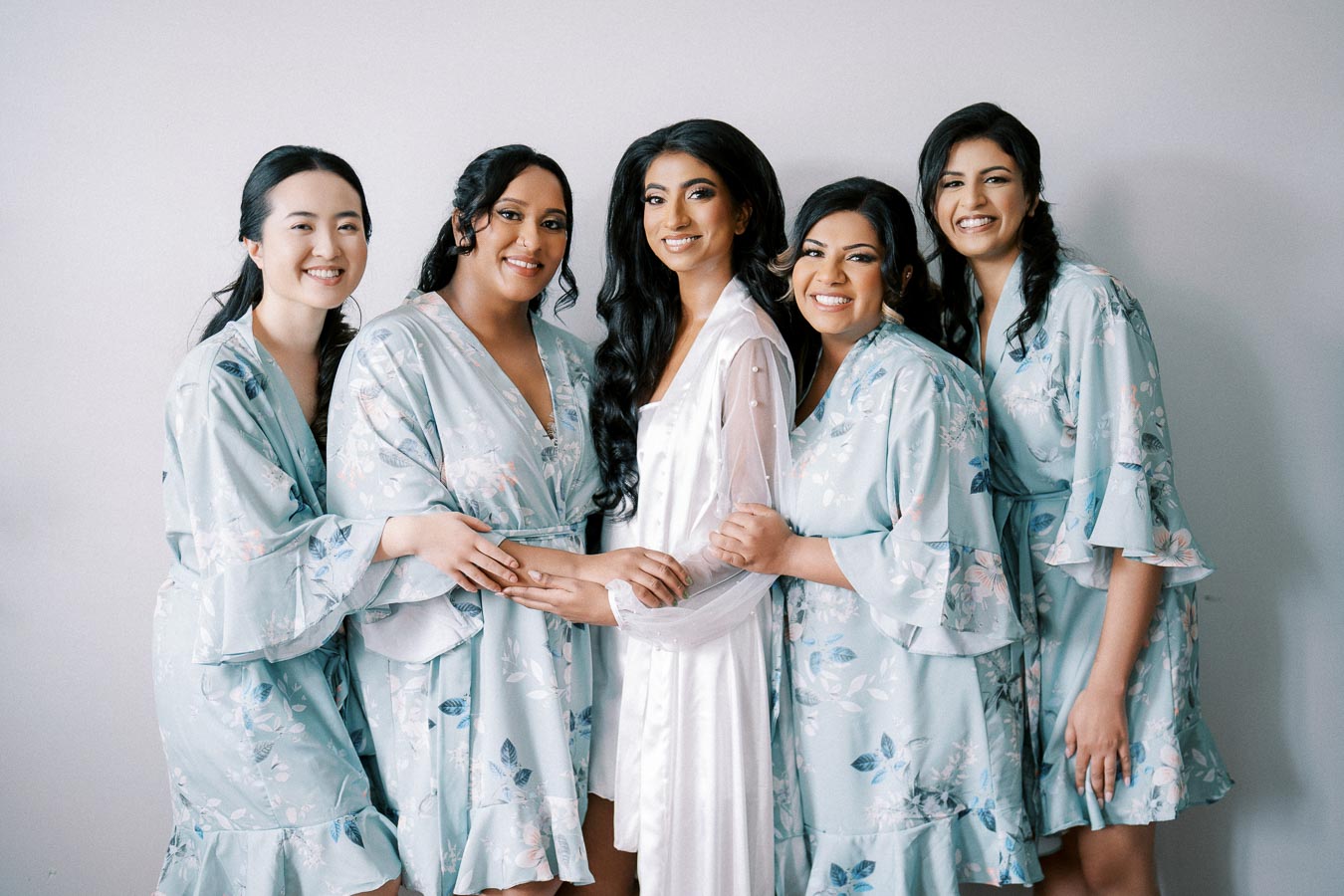 A bridal party of five women wearing floral robes gather for a pre-wedding photoshoot, with the bride in the center wearing a white robe, radiating joy and togetherness.