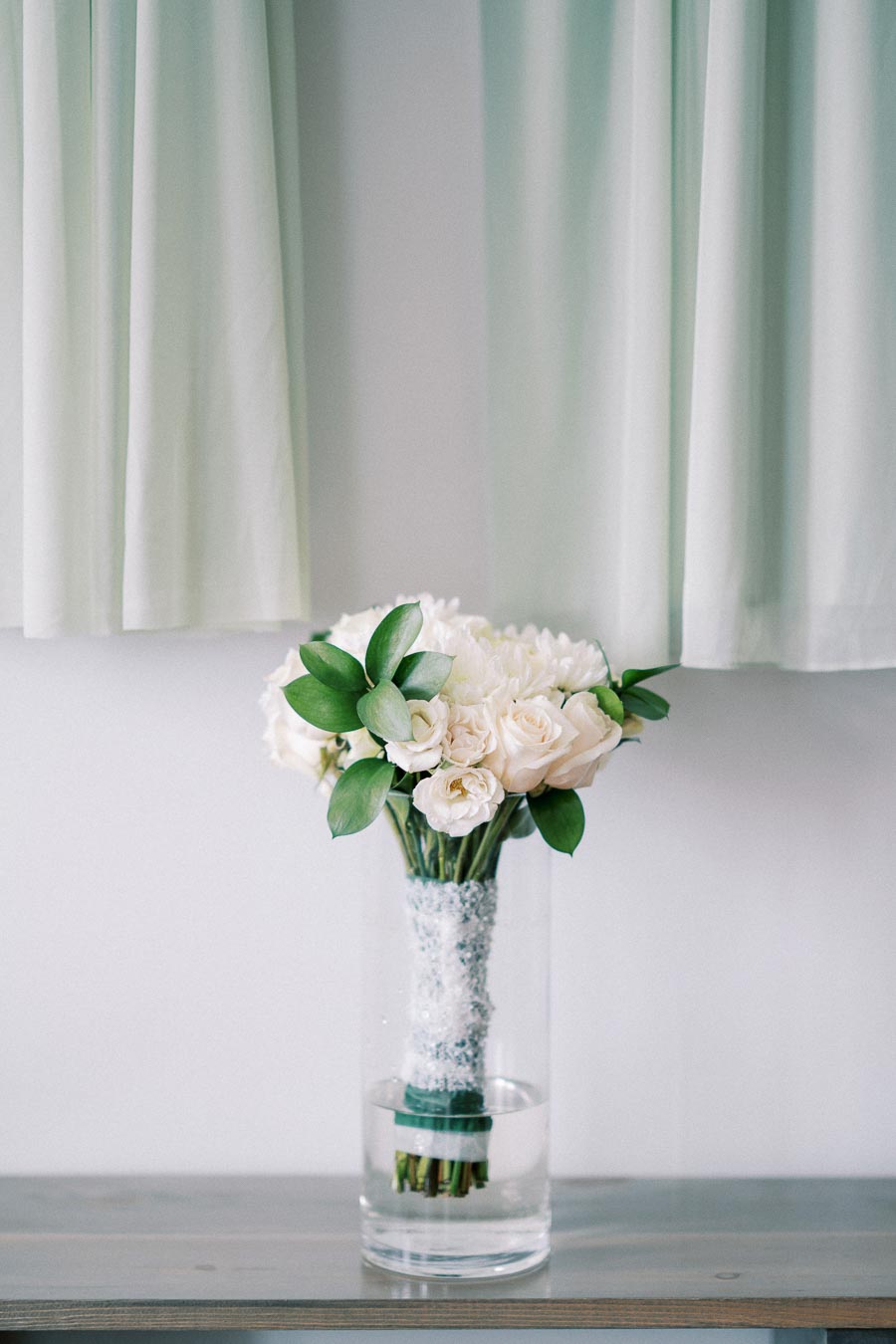 A beautiful bouquet of white roses and greenery in a clear vase, set against a soft pastel background with light curtains.