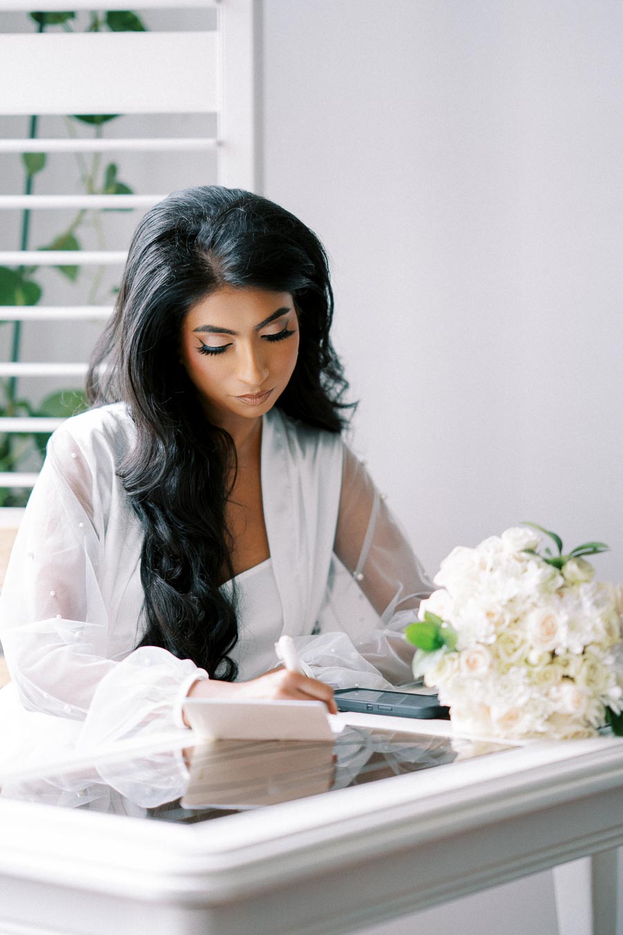 Elegant woman in a white robe writing in a notebook at a glass table, with a bouquet of white flowers nearby, set in a softly lit room.