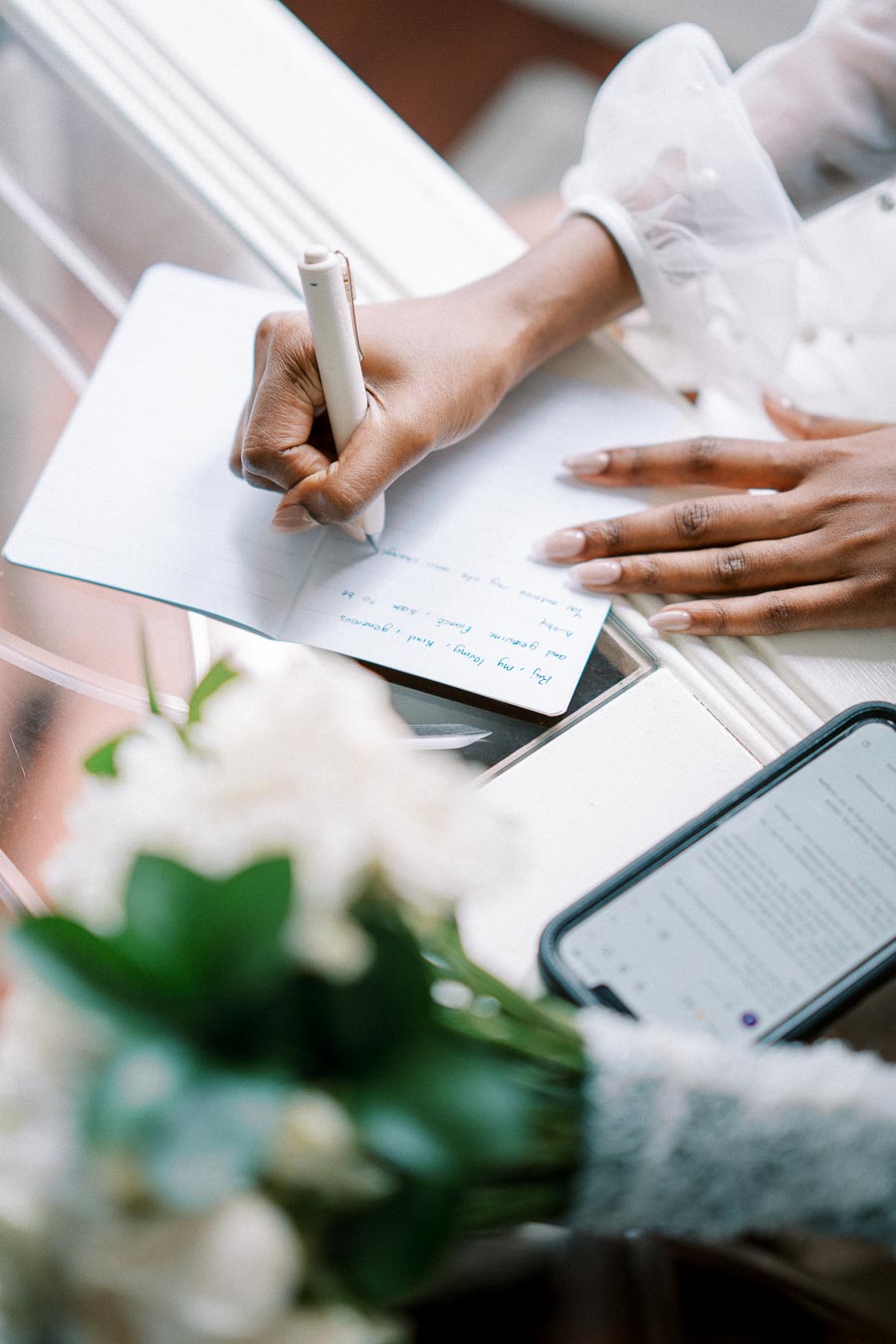 A person writing in a notebook with a white pen on a glass table, accompanied by a smartphone and a bouquet of white flowers in the foreground.