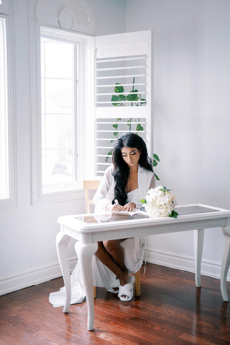 A woman in a white robe writing at a small white desk by a window, with a bouquet of white flowers in a bright and airy room.