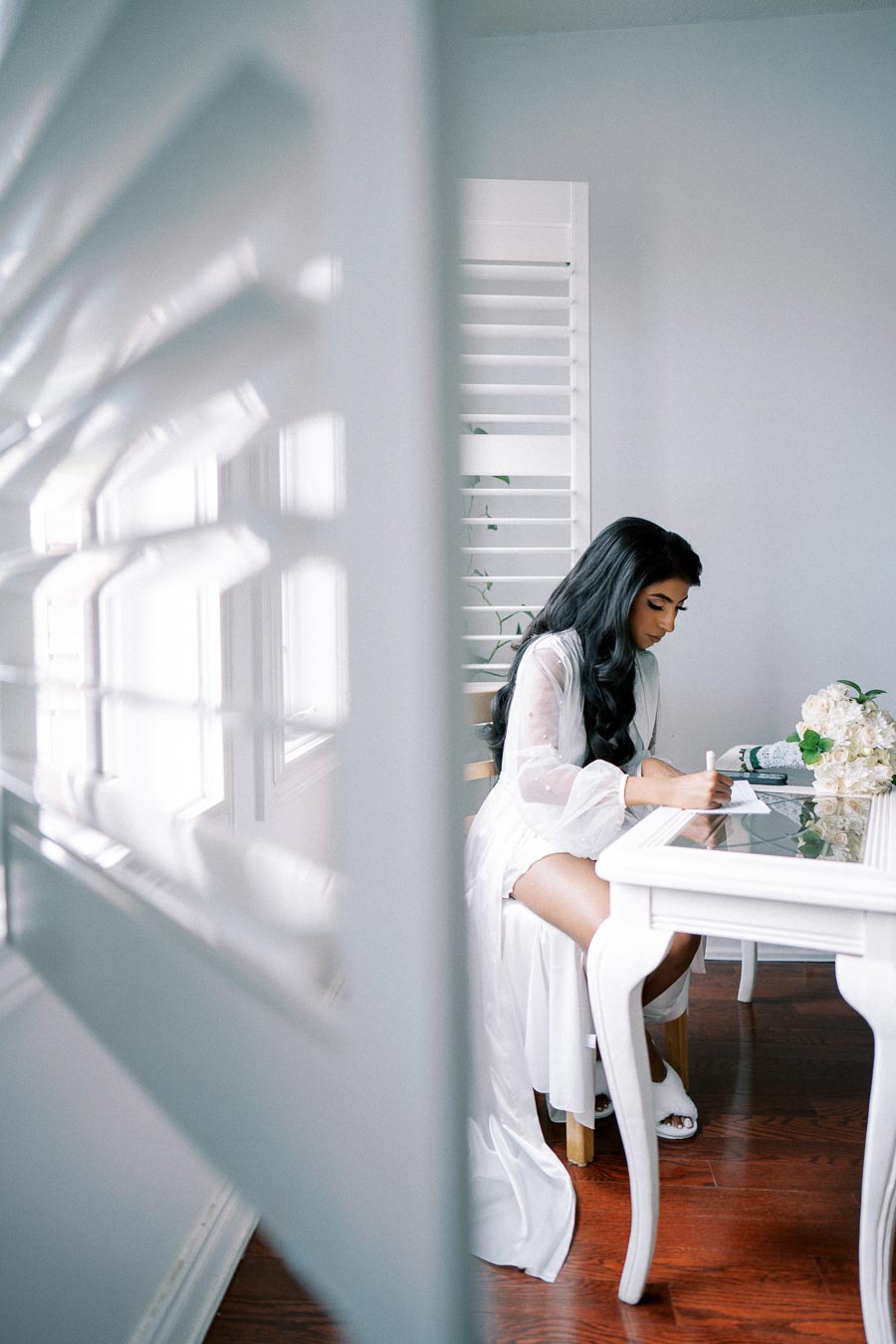 Young woman in white robe writing at a glass-top table, with soft window lighting filtering through blinds, and a bouquet of white flowers nearby.