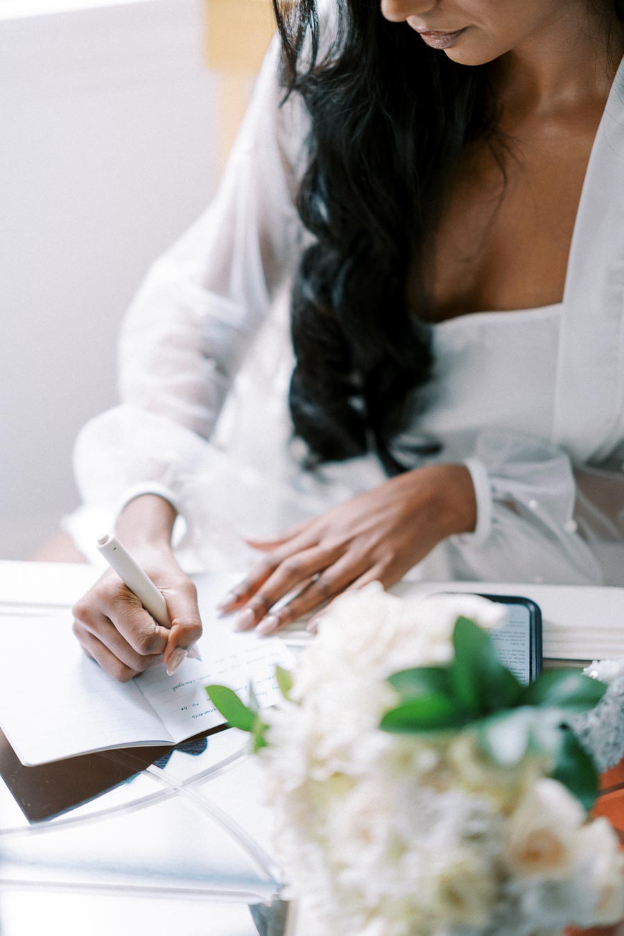 A woman in a sheer white robe writing in a notebook at a desk, with a smartphone nearby and a bouquet of white flowers in the foreground.