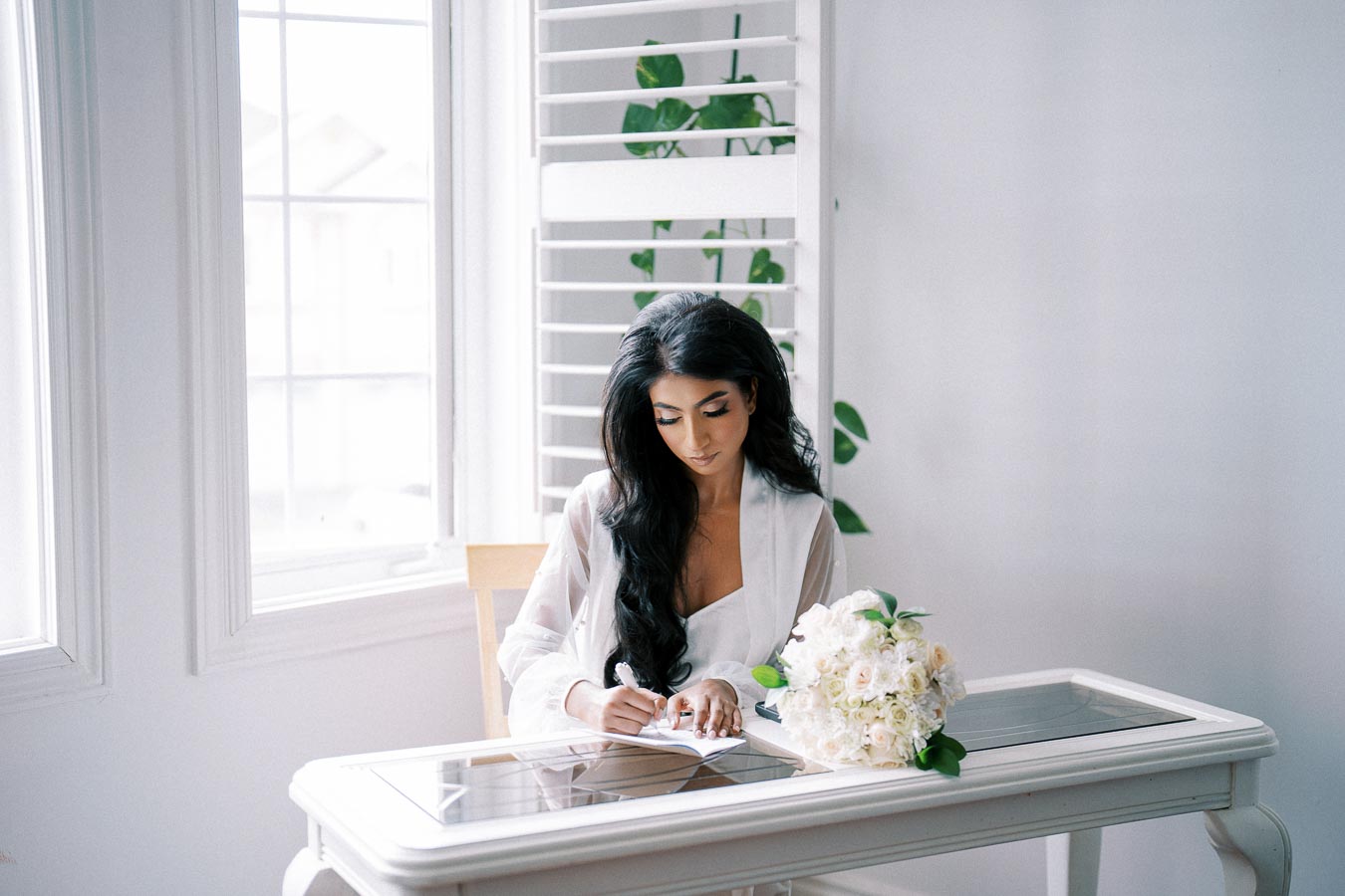 A woman in a white robe writing on a notepad at a white desk with a bouquet of flowers, set in a bright room with large windows and white shutters.