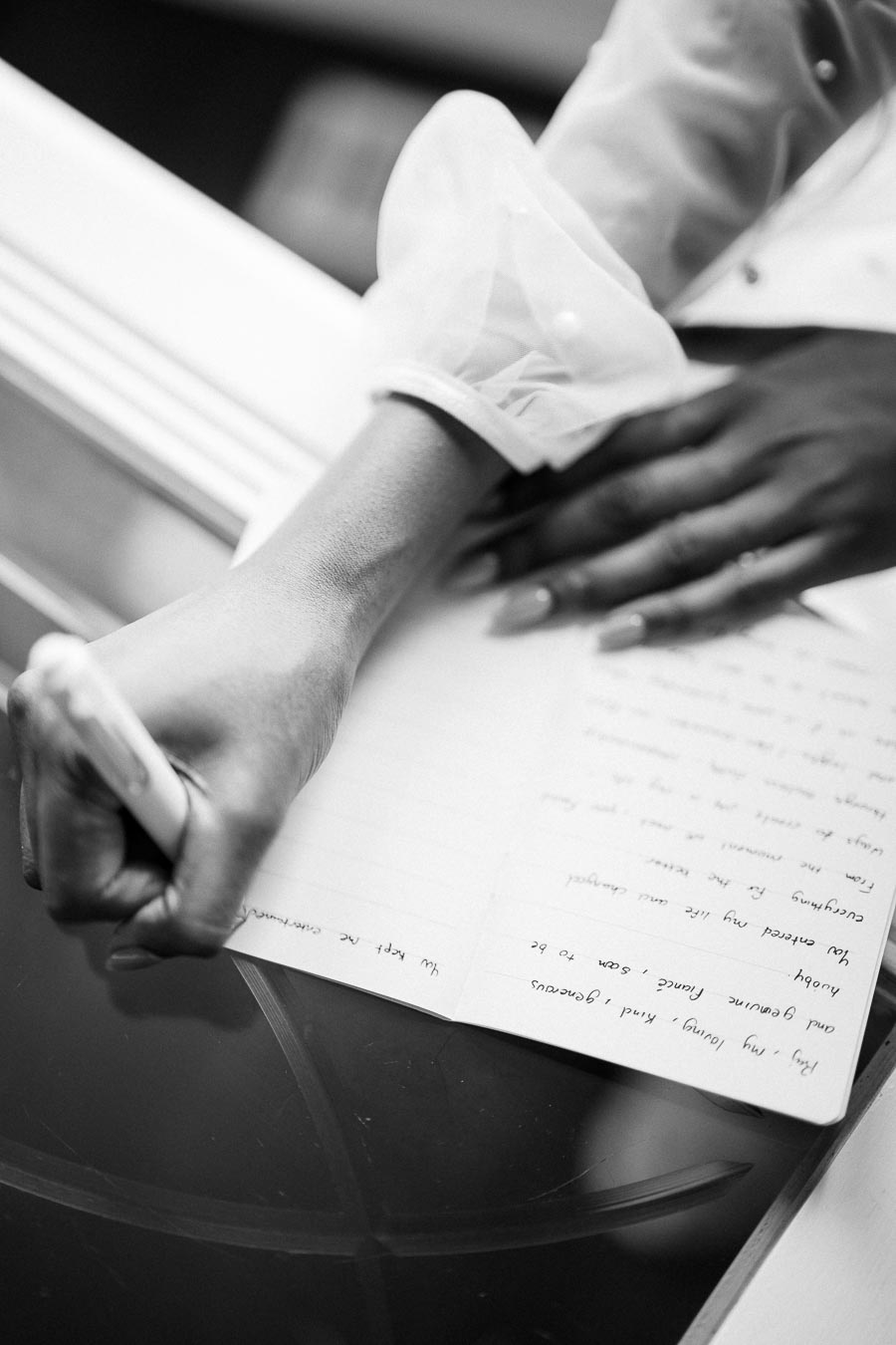 Black and white image of a person writing in a journal with a pen, wearing a sheer-sleeved blouse, highlighting creativity and personal reflection.