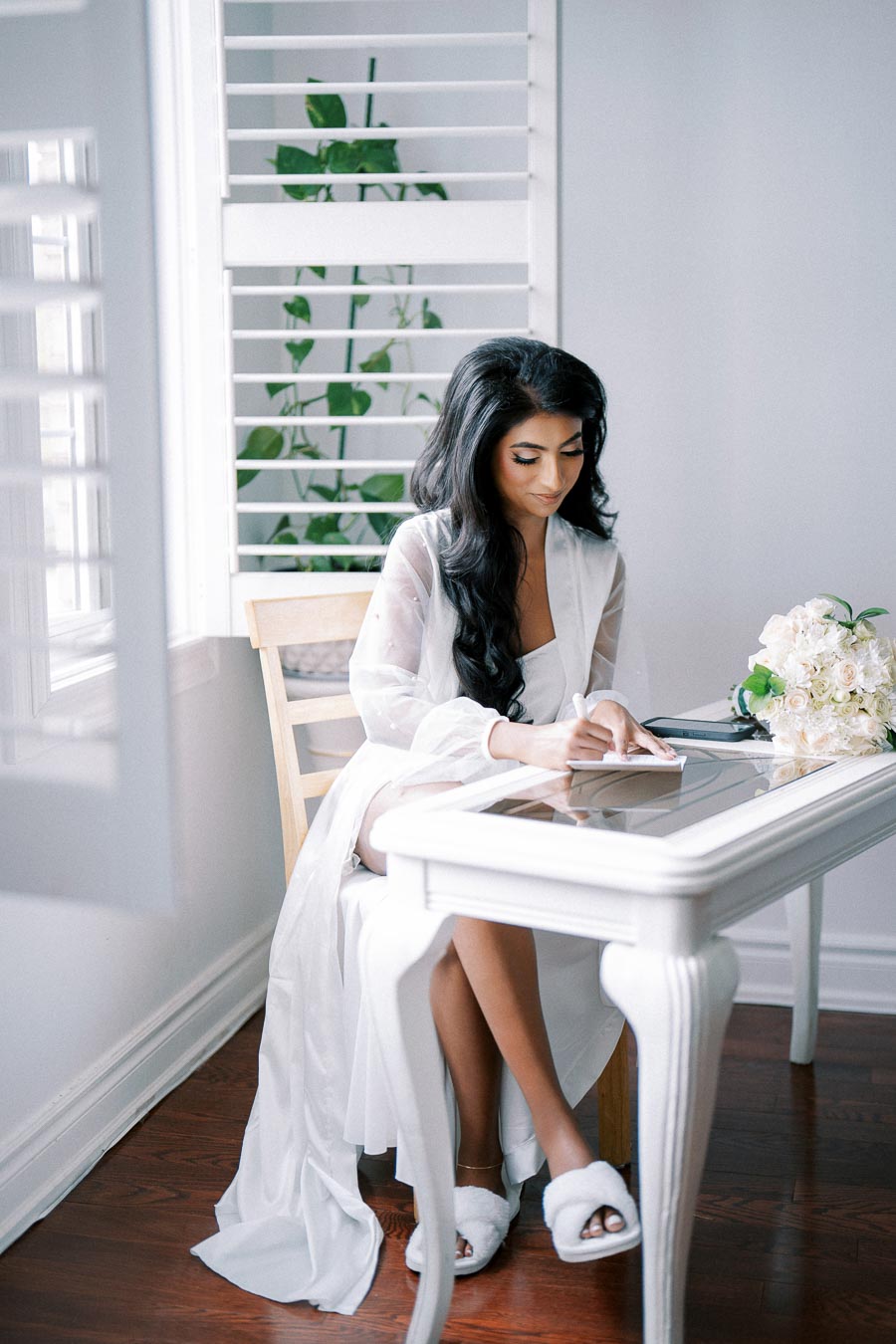 A woman in a white robe writes a note at a glass-topped desk next to a bouquet of white flowers, in a bright room with wooden floors and shuttered windows.