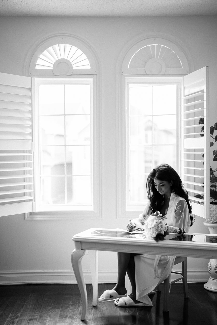 Black and white image of a woman in robe sitting at a table by large windows, writing with a bouquet of flowers on the table, creating a serene atmosphere.