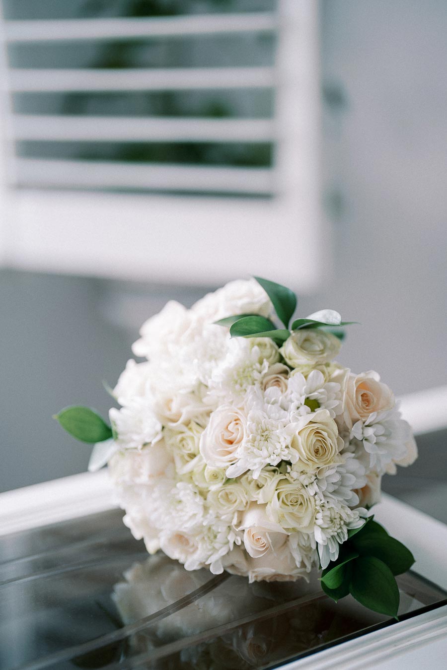 A beautiful bridal bouquet featuring white roses and chrysanthemums with green leaves, elegantly arranged on a reflective surface, perfect for a wedding or elegant event.