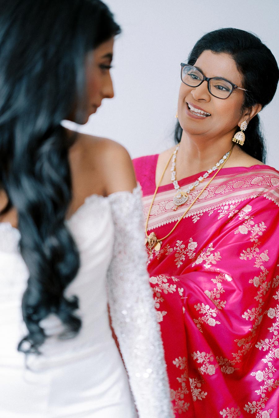A bride in a white gown shares a joyful moment with a woman in a vibrant pink sari with golden embroidery, featuring elegant jewelry, in a well-lit room.