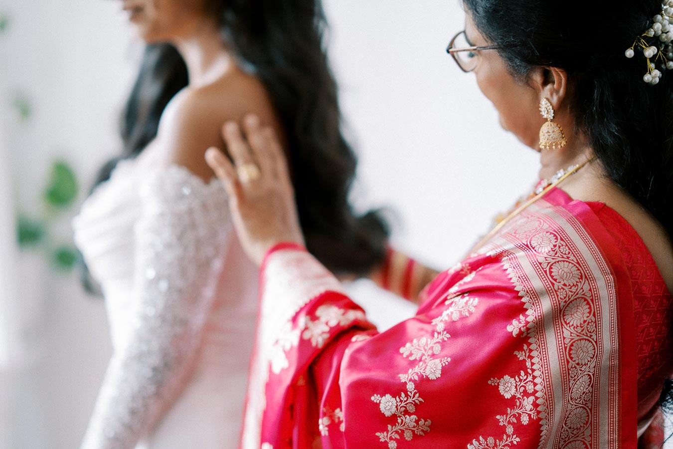A woman in traditional attire gently adjusts the bridal gown of a bride, showcasing cultural elegance and intimate bonding moments before a wedding ceremony.