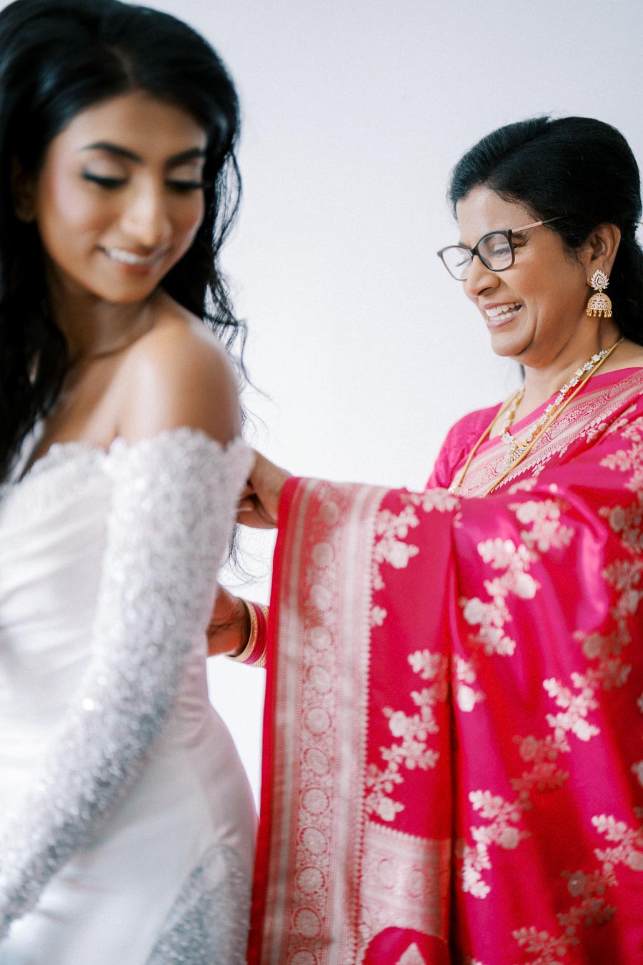 A bride getting ready with the help of her mother, who is wearing a vibrant pink and gold saree, showcasing a touching family moment before the wedding ceremony.