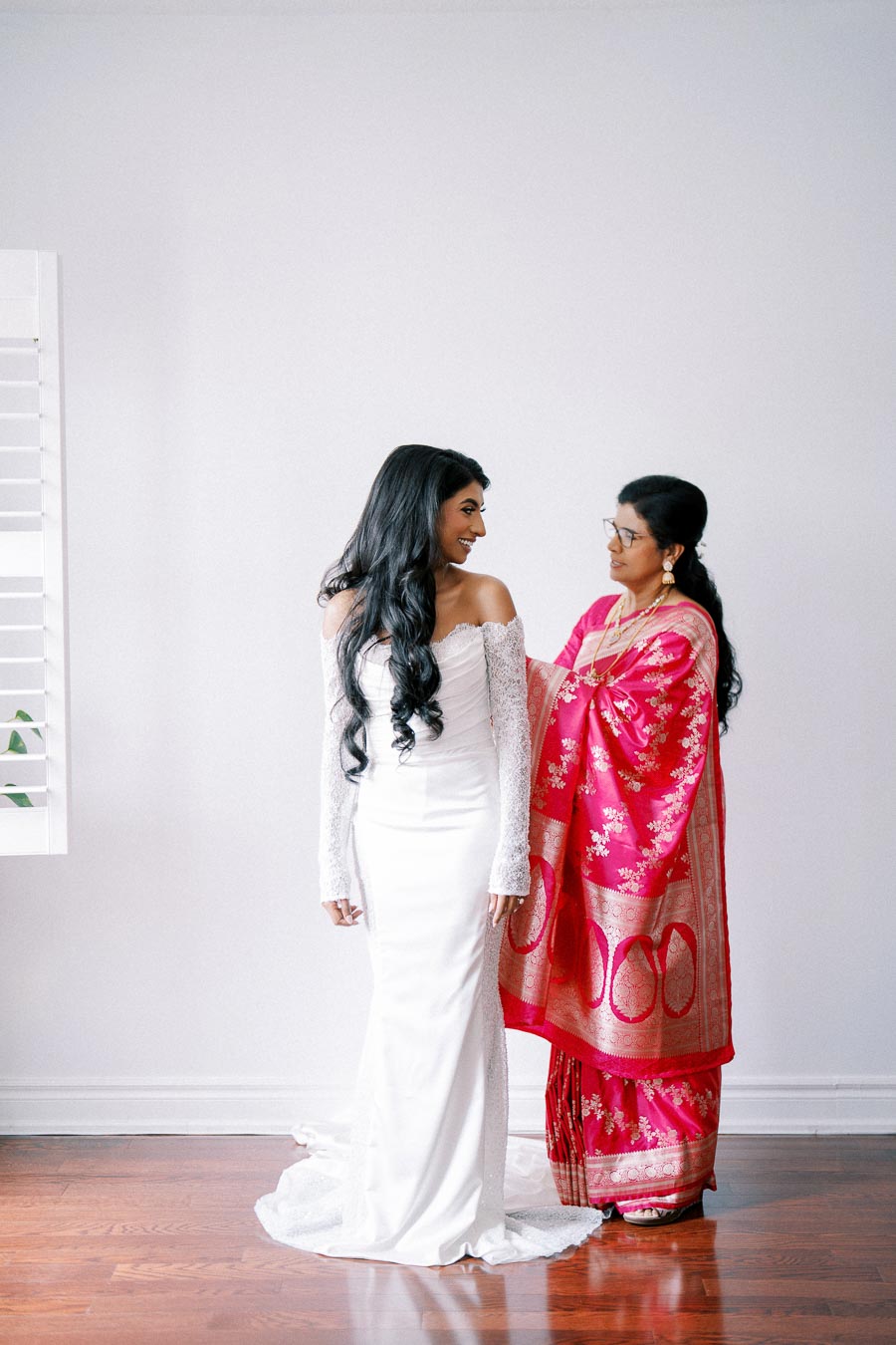 A bride in a white wedding dress smiles at a woman in a vibrant red and gold saree, preparing for a wedding ceremony.