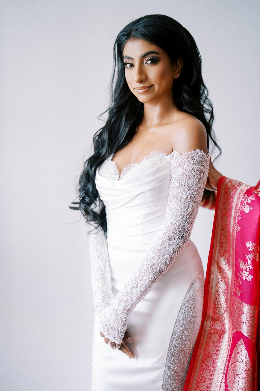 Elegant bride in a white wedding gown with lace sleeves, smiling softly against a light background. A pink and gold embroidery detail can be seen to the side, adding a touch of traditional color.