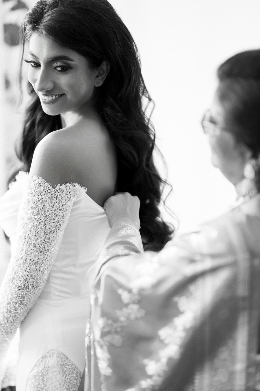 Bride getting ready in elegant lace wedding dress, assisted by another person, smiling over her shoulder in a beautifully lit room.