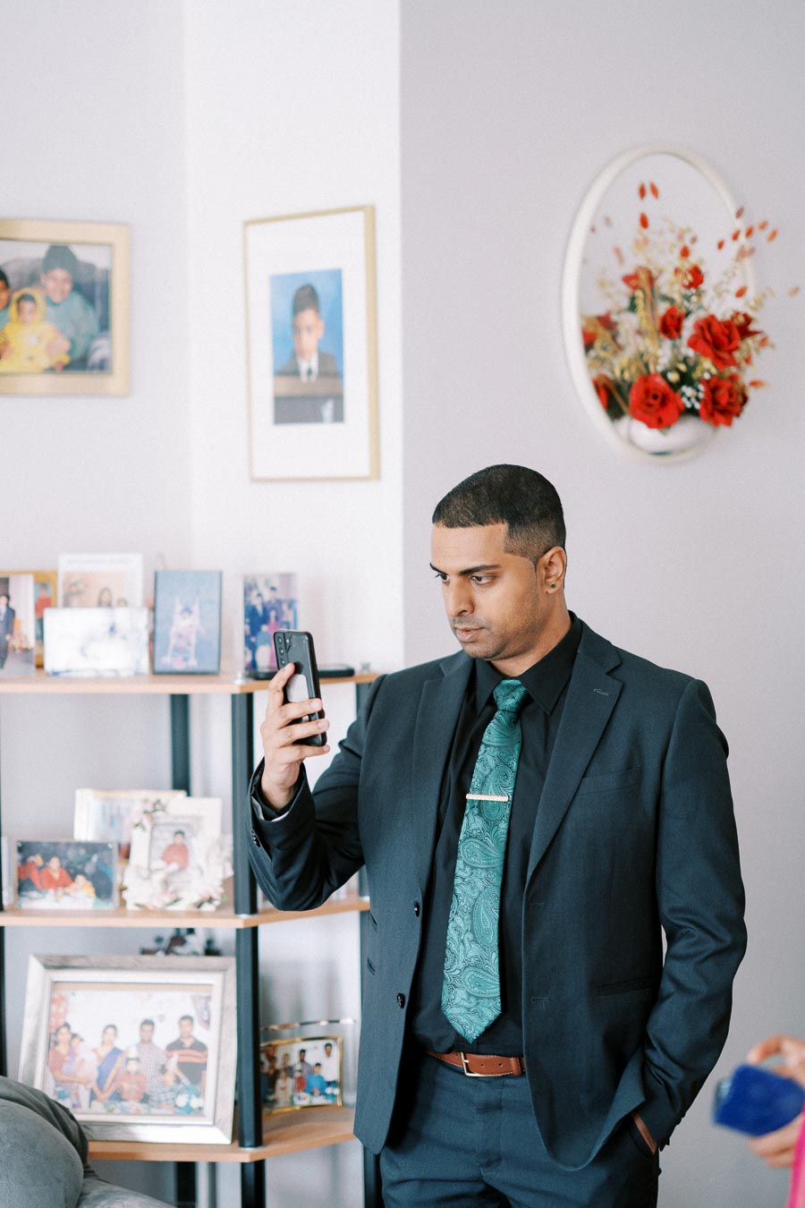 Man in a suit holding a smartphone in a living room with family photos and decorative wall art.
