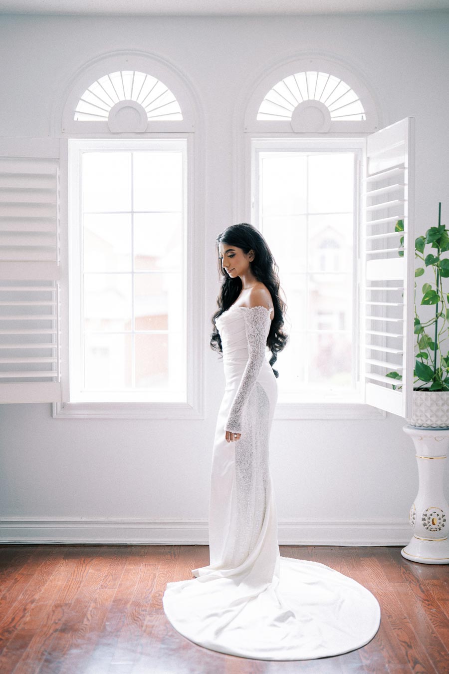 Bridal portrait of a woman in an elegant, long white wedding dress with lace sleeves, standing gracefully in a bright room with large windows and wooden floors.