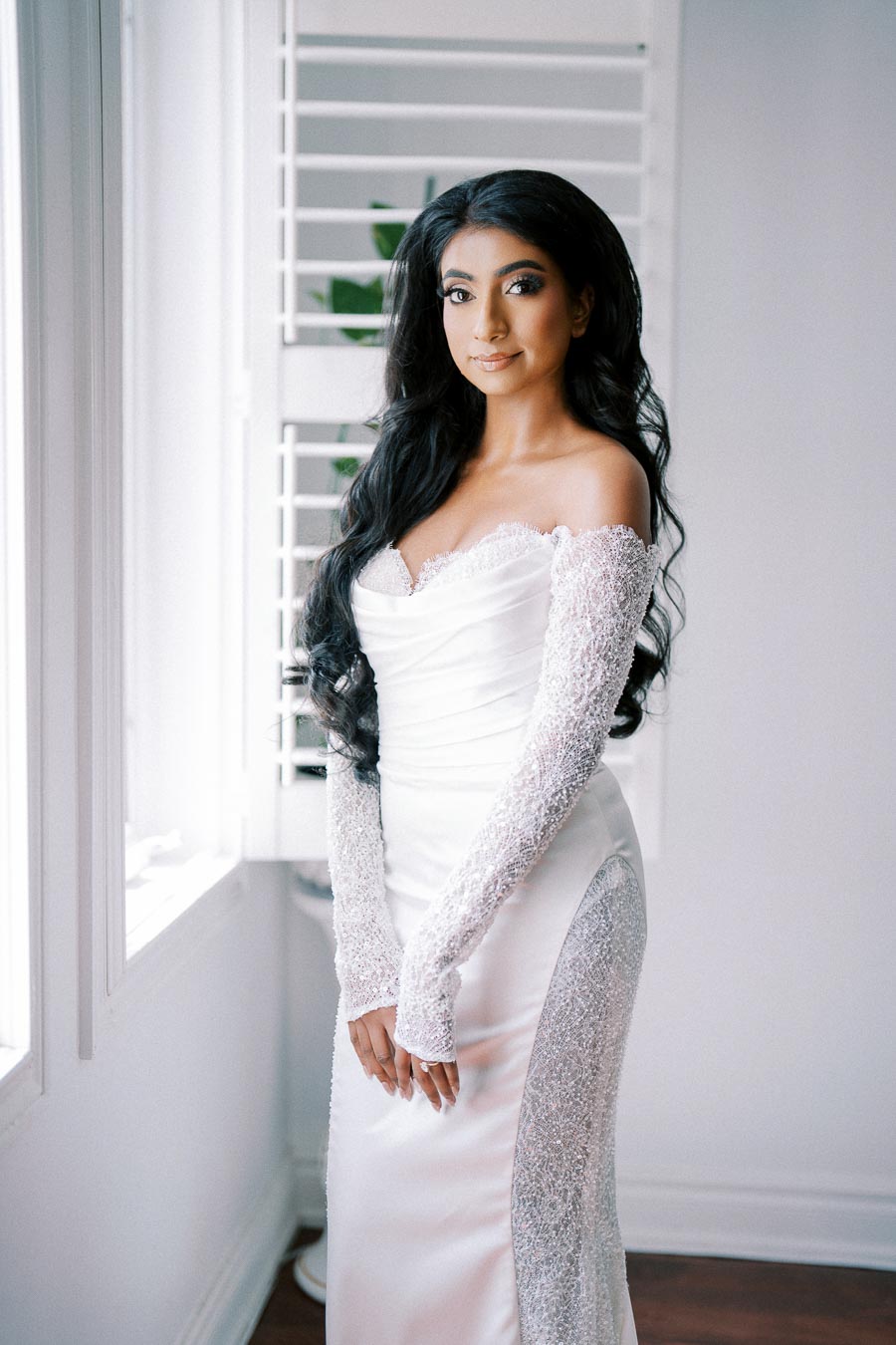 A bride in a white, off-the-shoulder wedding dress with intricate lace sleeves, standing by a window.