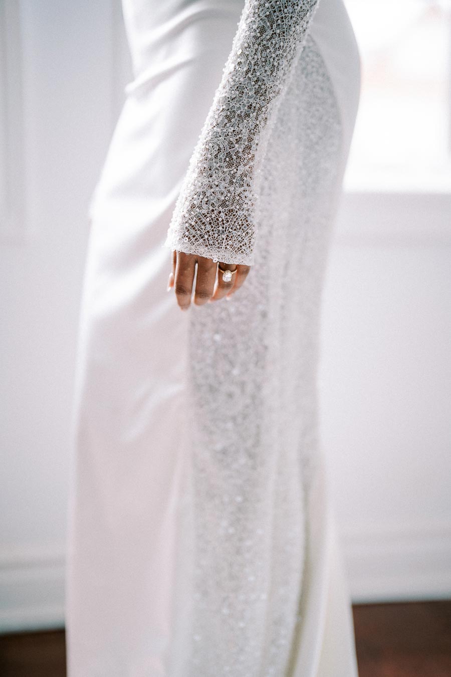 Close-up of a woman's hand adorned with an elegant, sparkling ring, featuring a stunning white gown with intricate lace details on the sleeve.