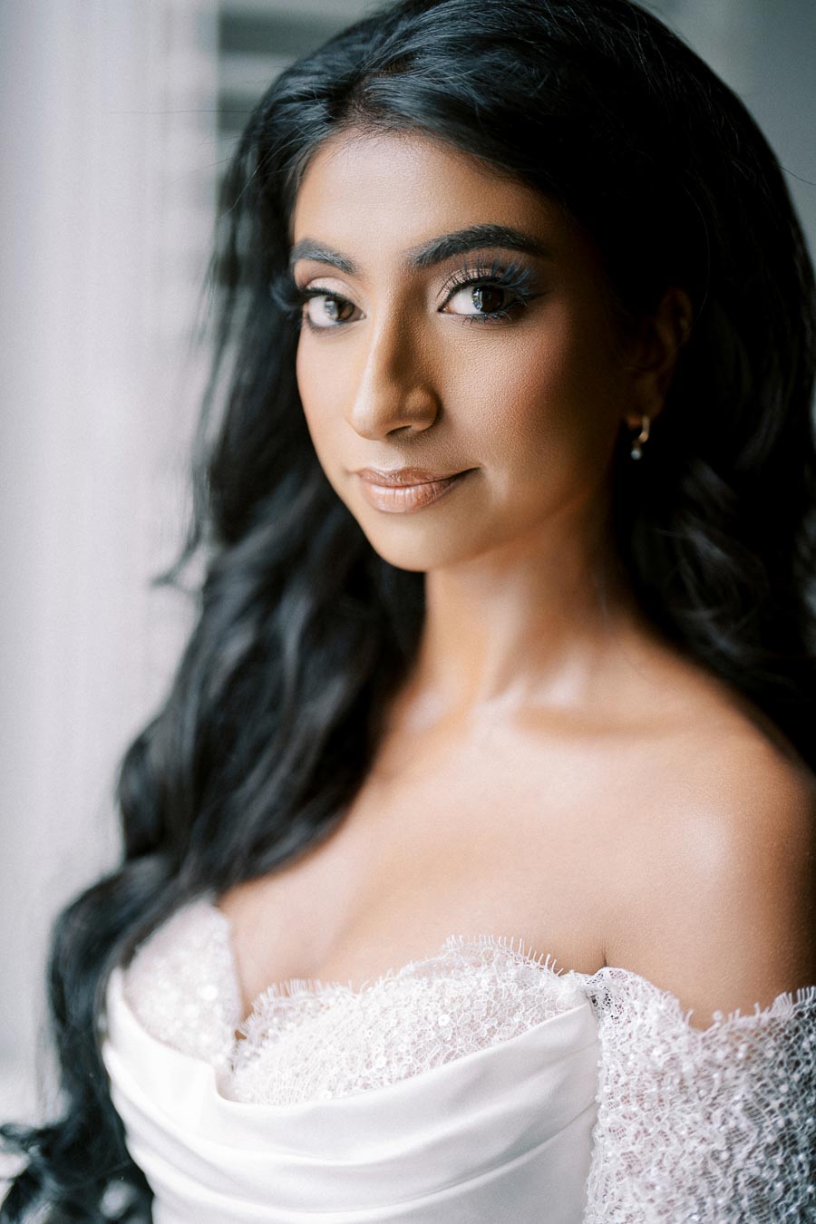 A bride with long black hair and elegant makeup, wearing an off-the-shoulder lace wedding dress, gazes softly towards the camera in natural light.