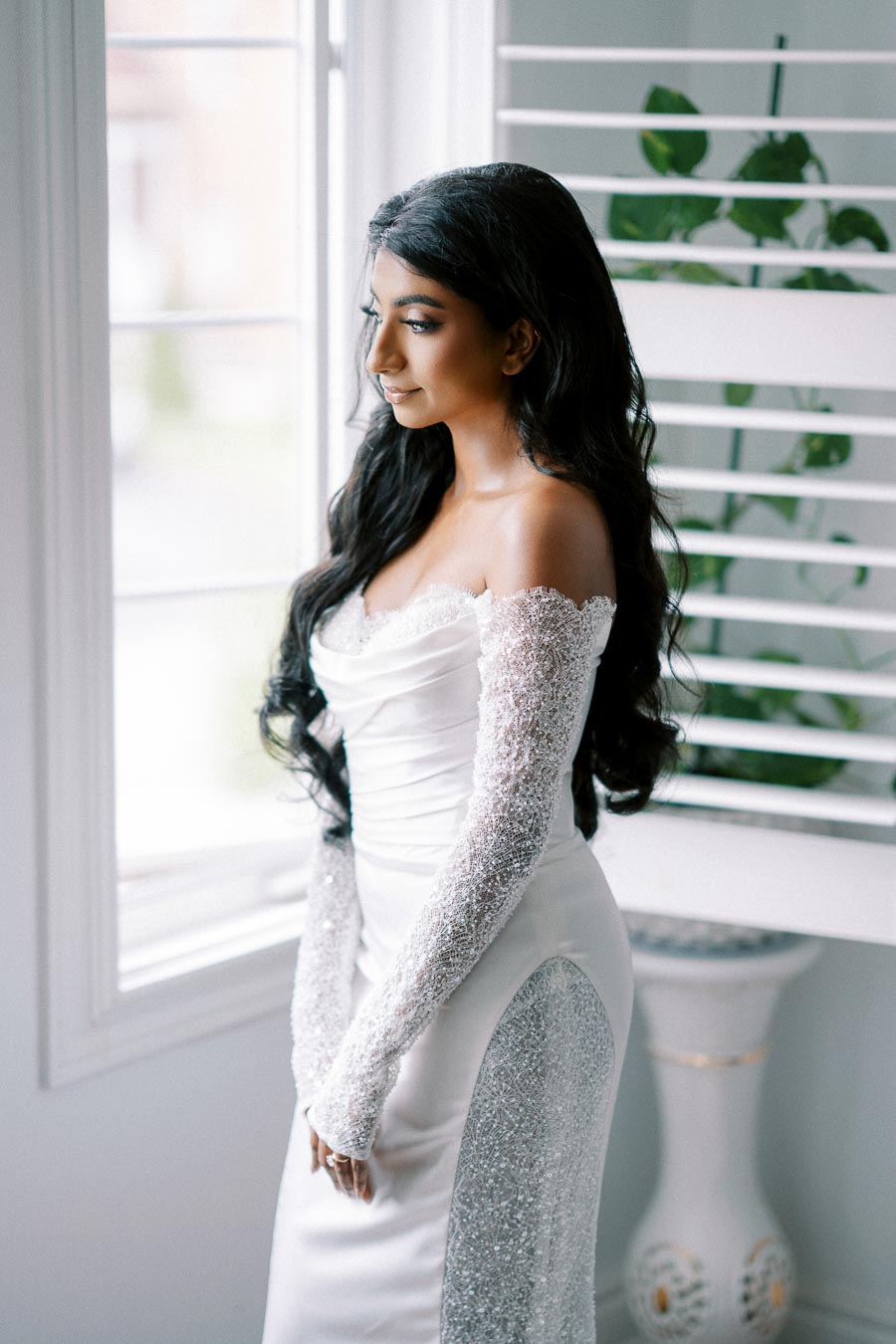 A woman in an elegant white wedding dress with intricate lace sleeves stands near a window with sunlight streaming in, complementing her long dark hair.